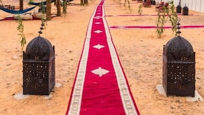 Un sentiero nel deserto con una moquette rossa che conduce a un edificio bianco, affiancato da palme e lanterne.