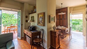 Uma sala de estar ampla com uma mesa de jantar, um sofá e uma escrivaninha com um computador. Há quadros nas paredes e uma janela com vista para o verde lá fora.