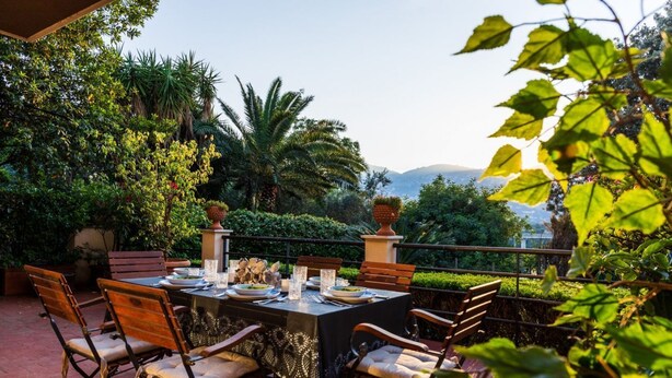 Uma área de refeições ao ar livre com uma mesa posta para quatro pessoas, rodeada por cadeiras, em um jardim exuberante com diversas plantas e vista para colinas distantes.
