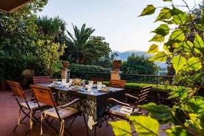 Ein Essbereich im Freien mit einem Tisch für vier Personen, umgeben von Stühlen, in einem üppigen Garten mit verschiedenen Pflanzen und Blick auf die entfernten Hügel.