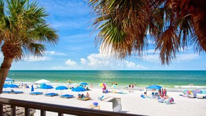 A beach with palm trees, umbrellas, and people. - Beachfront Balcony Corner Unit on Madeira Beach. Updated, Comfortable 1st Floor, with Pool & Grill. (Madeira Beach)