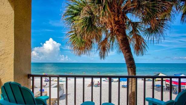 A balcony with turquoise chairs overlooking a beach with umbrellas and people. - Beachfront Balcony Corner Unit on Madeira Beach. Updated, Comfortable 1st Floor, with Pool & Grill. (Madeira Beach)