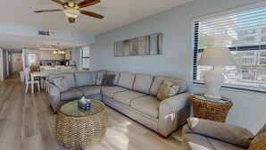 A modern living room with a sectional sofa, a round woven coffee table, and a floor lamp. - Beachfront Balcony Corner Unit on Madeira Beach. Updated, Comfortable 1st Floor, with Pool & Grill. (Madeira Beach)