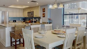 A modern kitchen with white cabinetry, a central island, and a dining area with white chairs and a table set for a meal. - Beachfront Balcony Corner Unit on Madeira Beach. Updated, Comfortable 1st Floor, with Pool & Grill. (Madeira Beach)