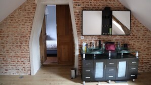 A room with a sloped ceiling, exposed brick walls, a wooden door, a black vanity with a glass sink, and a mirror above it. - Authentic Norman house between countryside and sea (Lindebeuf)
