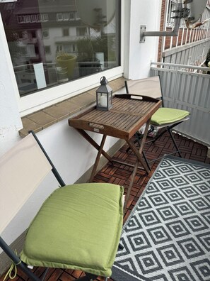 Un balcón con una mesa de madera, dos sillas con cojines verdes, un farol y una alfombra estampada.
