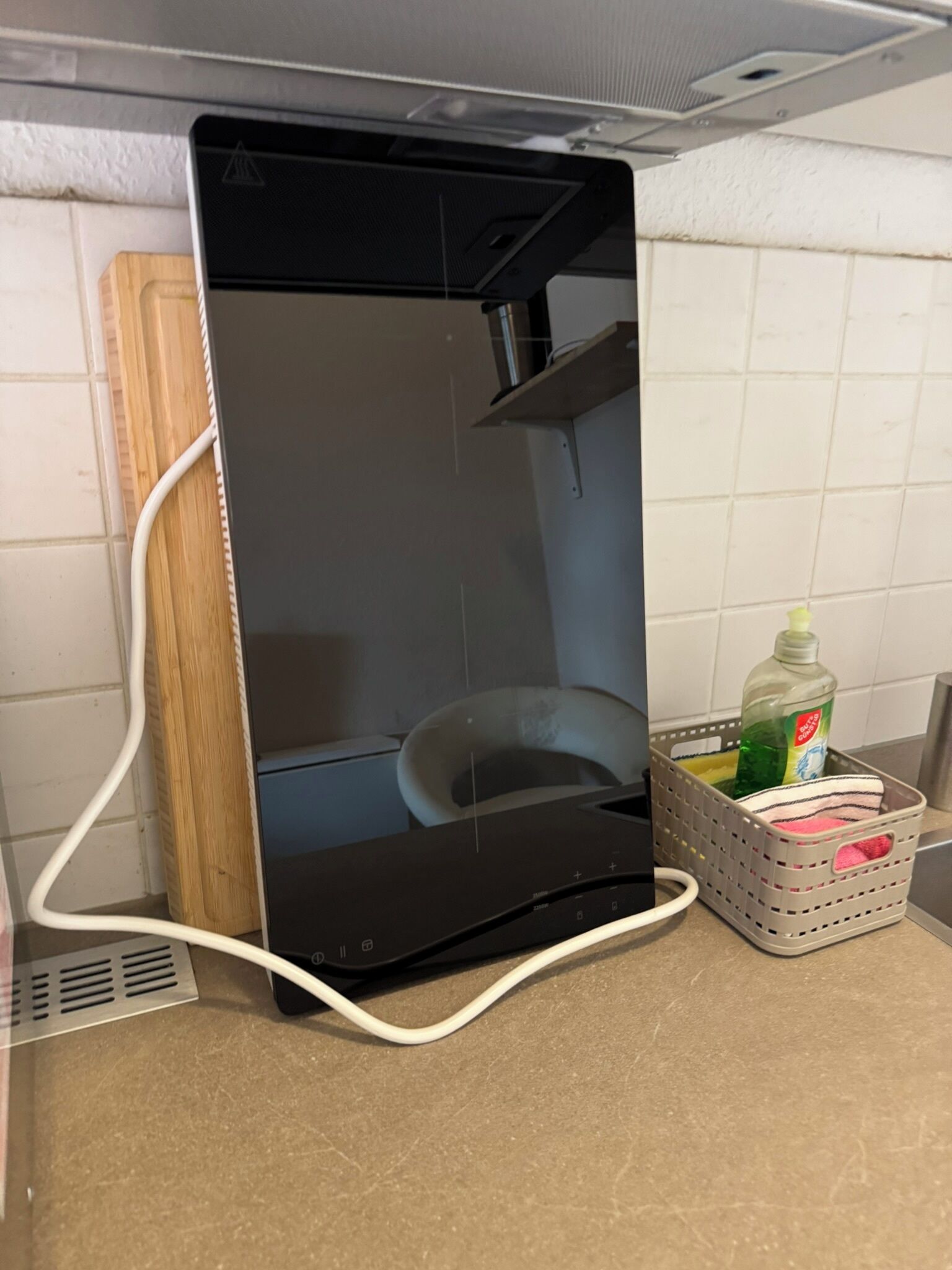 Una cocina con electrodoméstico negro, tabla de cortar de madera, una silla blanca y una cesta de ropa con detergente.