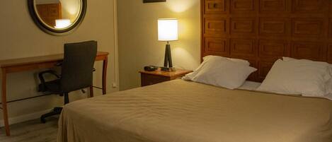 A hotel room with a bed, a desk with a chair, a nightstand with a lamp, a mirror, and a wooden headboard.