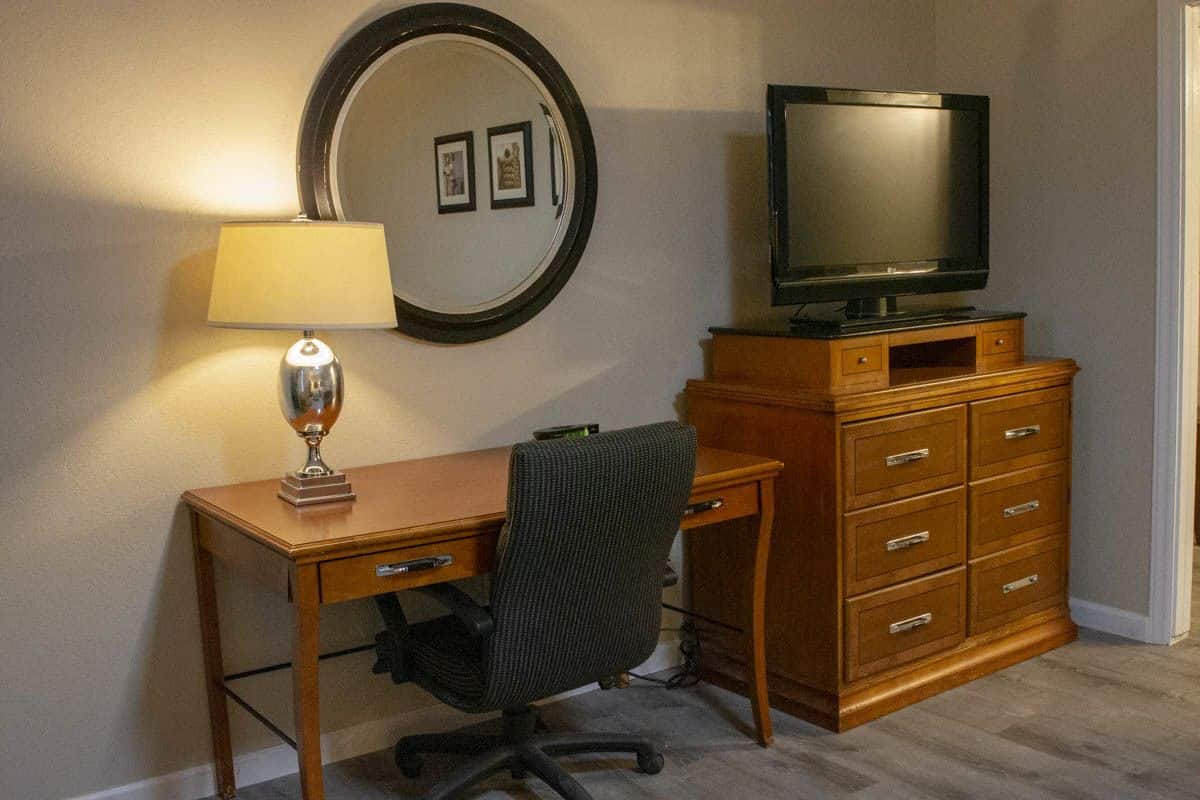 Quarto com escrivaninha de madeira, uma cadeira, televisão sobre aparador de madeira, luminária e espelho na parede.