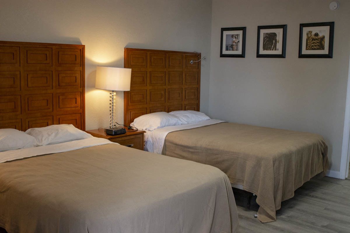 Habitación de hotel con dos camas, cabecero de madera, mesita de noche con lámpara y tres cuadros enmarcados en la pared.