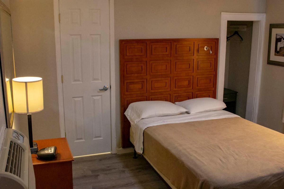 Una habitación de hotel con cabecera de madera, una cama con sábanas blancas, una lámpara de noche y una mesita con un control remoto.