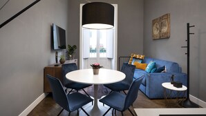 A modern living room with a blue sofa, a round dining table with chairs, a TV mounted on the wall, and a large window with curtains.