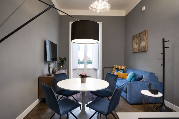 A modern living room with a blue sofa, a round dining table with chairs, a TV mounted on the wall, and a large window with curtains.