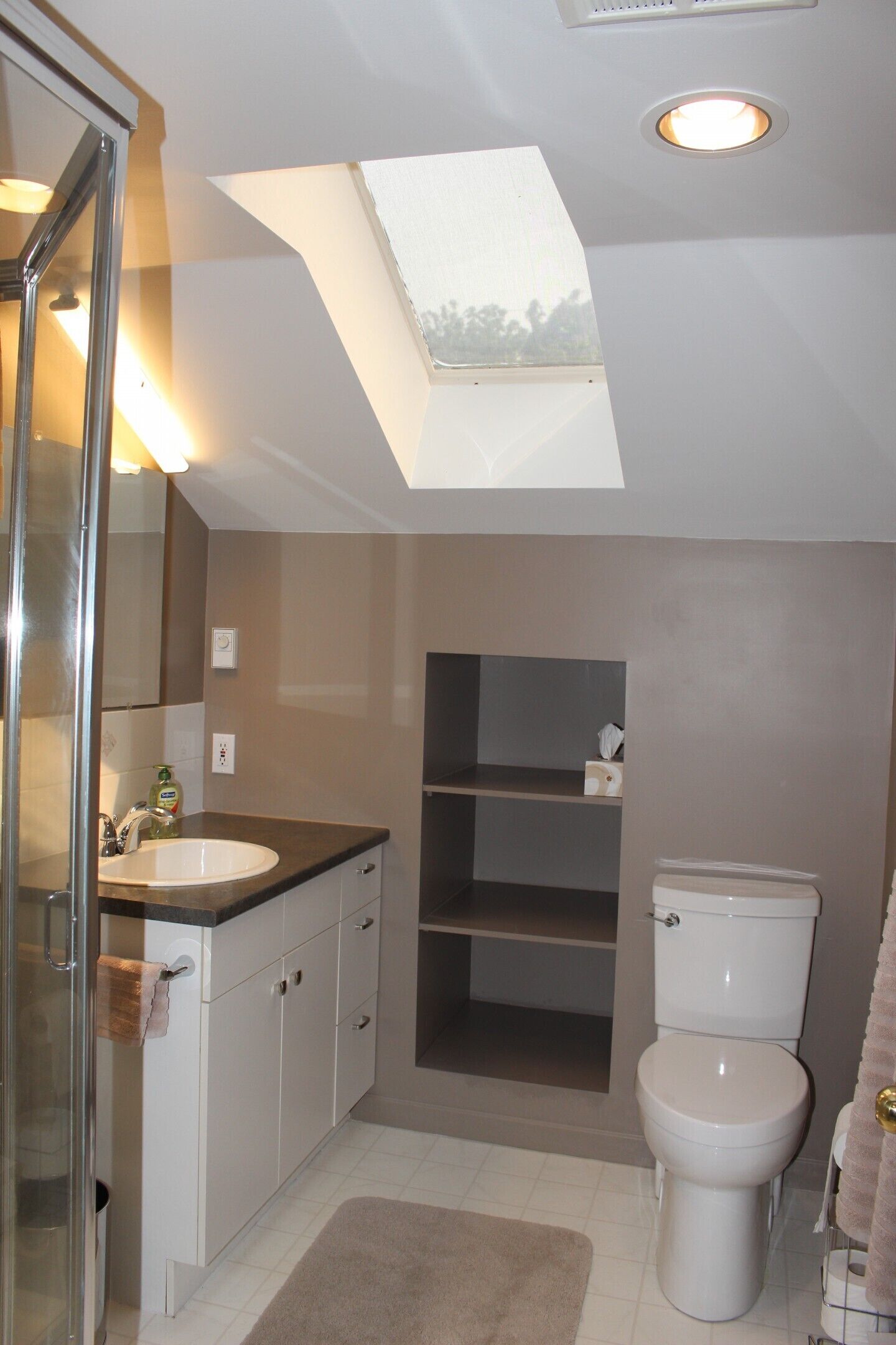 Een moderne badkamer met dakraam, een douchecabine van glas, een witte wastafel en een toilet.