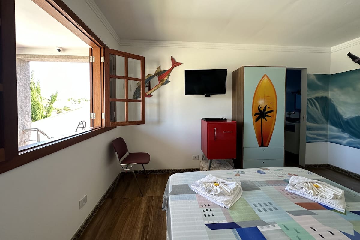 A hotel room with a bed, a red cabinet, a television, a window with a view of greenery, and a surfboard artwork on the wall.