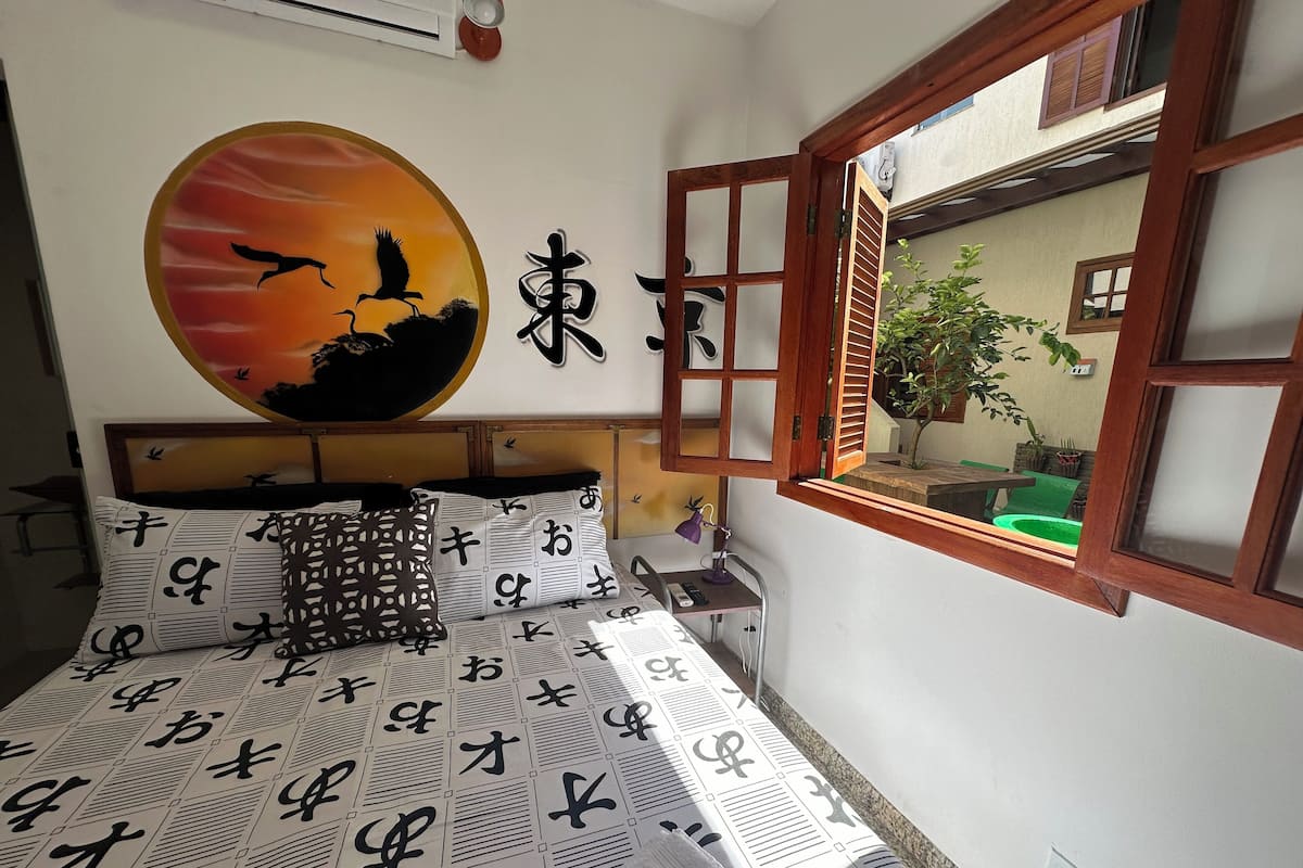 A bedroom with a bed, pillows with Japanese characters, a wall-mounted painting of birds, and a window with a view of the outside.
