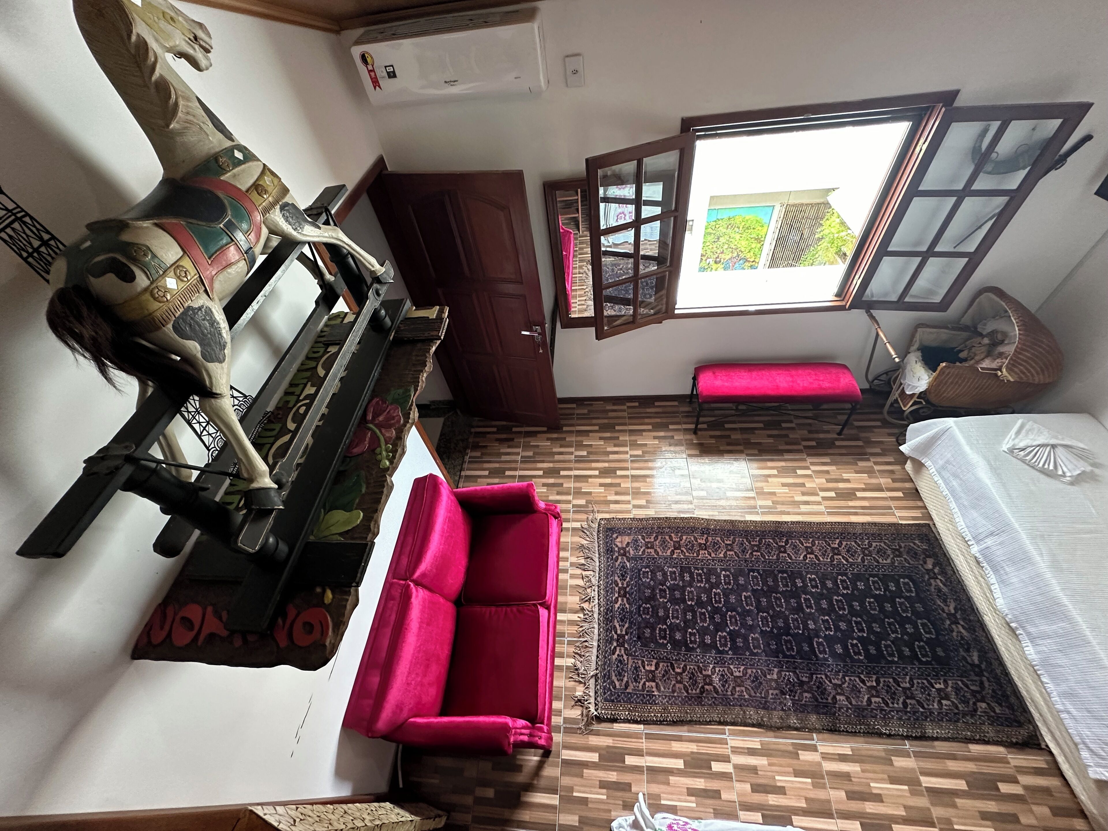 A compact room with a wooden floor, a red sofa, a patterned rug, a bed with a white sheet, a wooden door, and a window with a view of greenery.