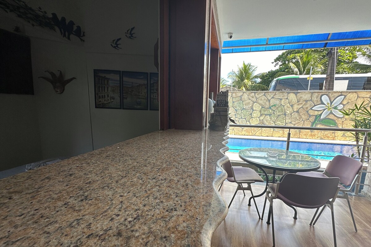A spacious indoor area with a granite countertop, a glass table, and chairs. There is a view of a swimming pool outside.