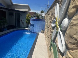 Una piscina con borde de azulejos azules, un muro de piedra con un diseño floral pintado, y un edificio con tejado de tejas al fondo.