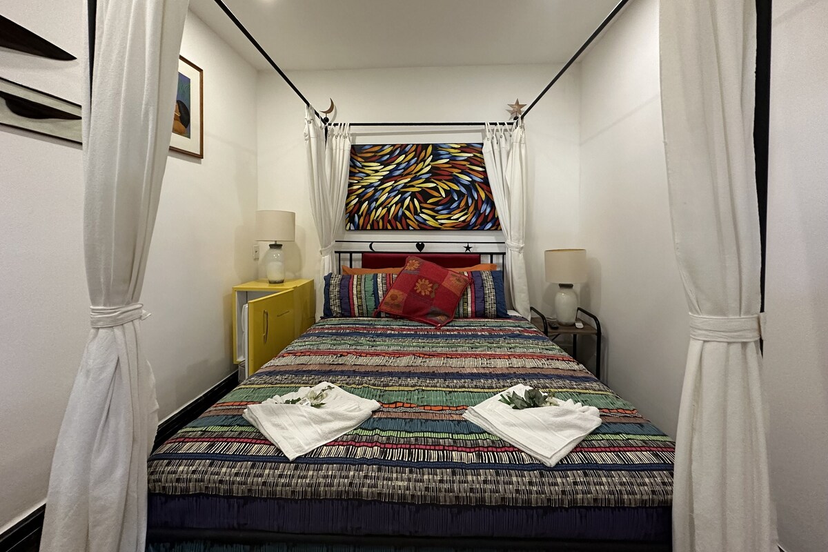 A bedroom with a bed, two bedside tables, a nightstand, a wall-mounted lamp, a ceiling light, and a window with curtains.