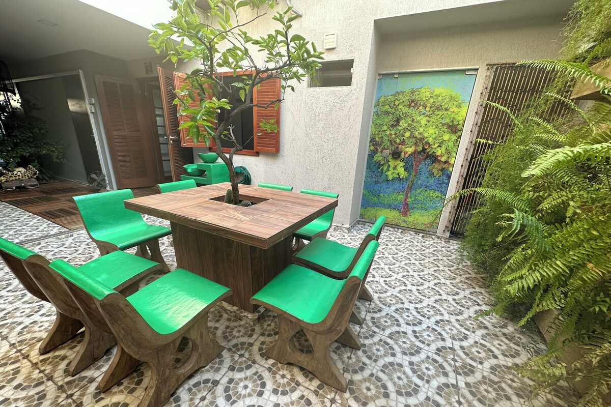 A patio with a wooden table and green chairs, surrounded by a mosaic-tiled floor and greenery.