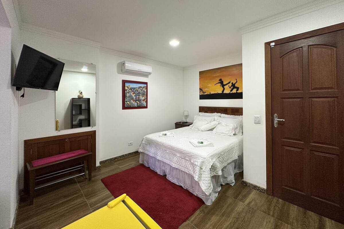 A bedroom with a bed, a TV mounted on the wall, a desk with a chair, a red rug, and a door.