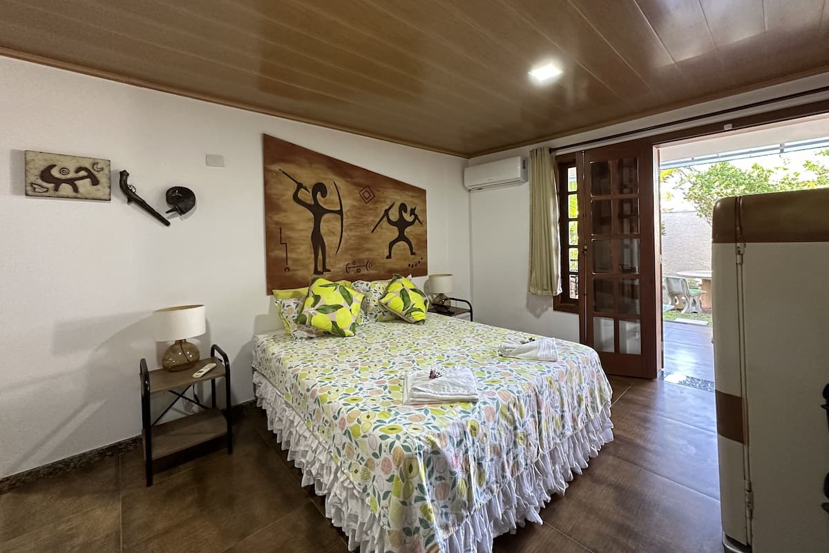 A bedroom with a wooden ceiling, a bed with a floral bedspread, a nightstand with a lamp, and artwork on the wall.