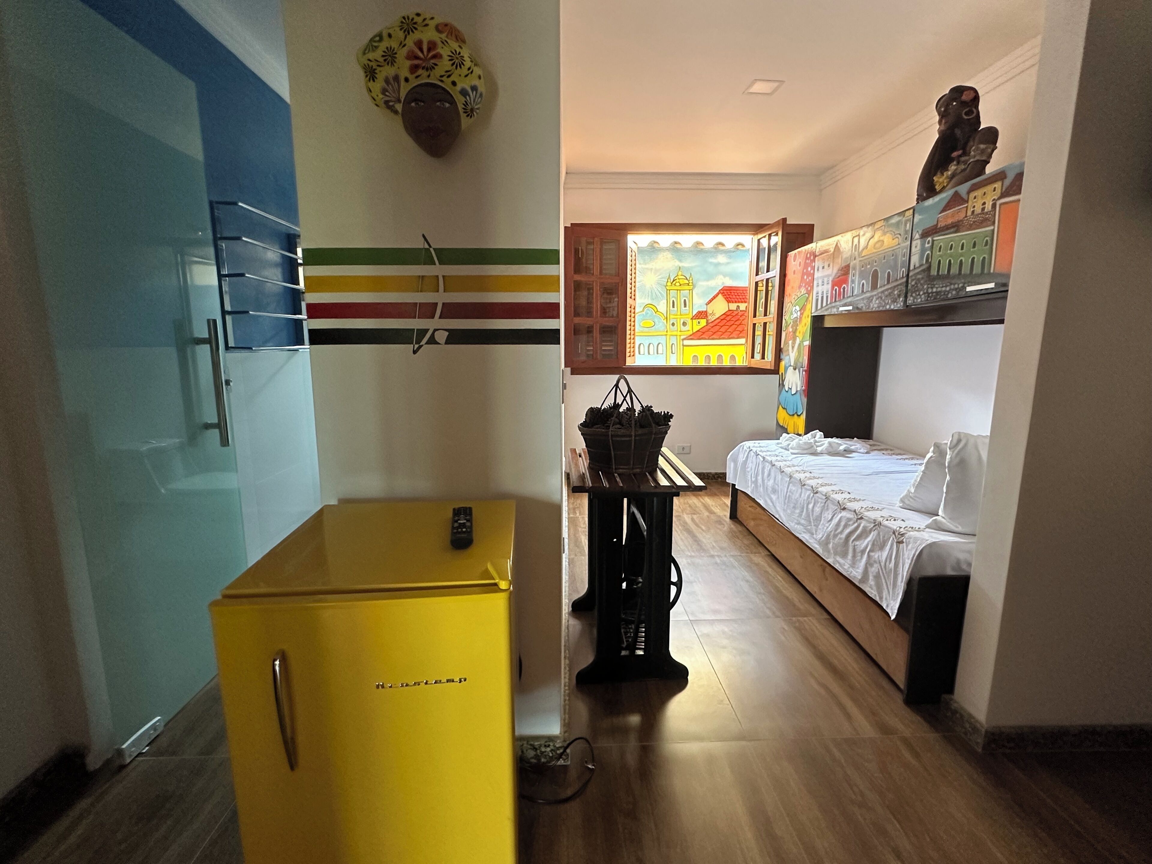 A compact hotel room with a bed, a small yellow refrigerator, a window with a view of buildings, and a wall-mounted shelf with decorative items.