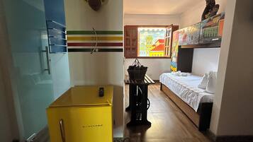 A compact hotel room with a bed, a small yellow refrigerator, a window with a view of buildings, and a wall-mounted shelf with decorative items.