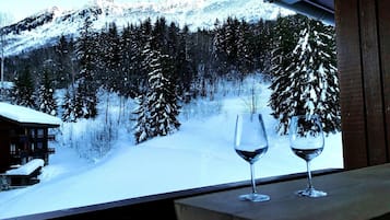 Une terrasse en bois avec deux verres à vin, donnant sur un paysage montagneux enneigé, avec de vastes forêts et un chalet.