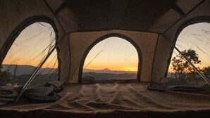 Una carpa con tres ventanas arqueadas, una cama en su interior y vistas a un atardecer sobre montañas.