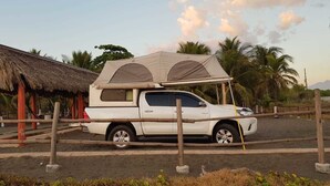 Una camioneta pickup blanca con techo de lona estacionada en un área de aparcamiento con una estructura de techo de paja y palmeras al fondo.