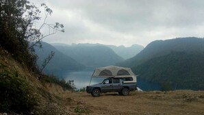 Un vehículo con una tienda de campaña en una ladera rocosa, con un lago y montañas al fondo.
