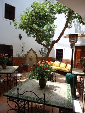 An outdoor patio with tables, chairs, a sofa, and a tree providing shade. - Riad Aldiana (Marrakech)