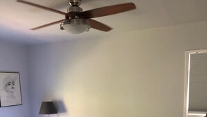 Ein ordentlich bezogenes Bett mit weißen Laken und einer dunkelgrauen Decke, ein Deckenventilator, eine Nachttischlampe und ein gerahmtes Bild an der Wand.