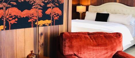 A hotel room with a large painting of trees, a wooden headboard, a white bed, and red velvet chairs.