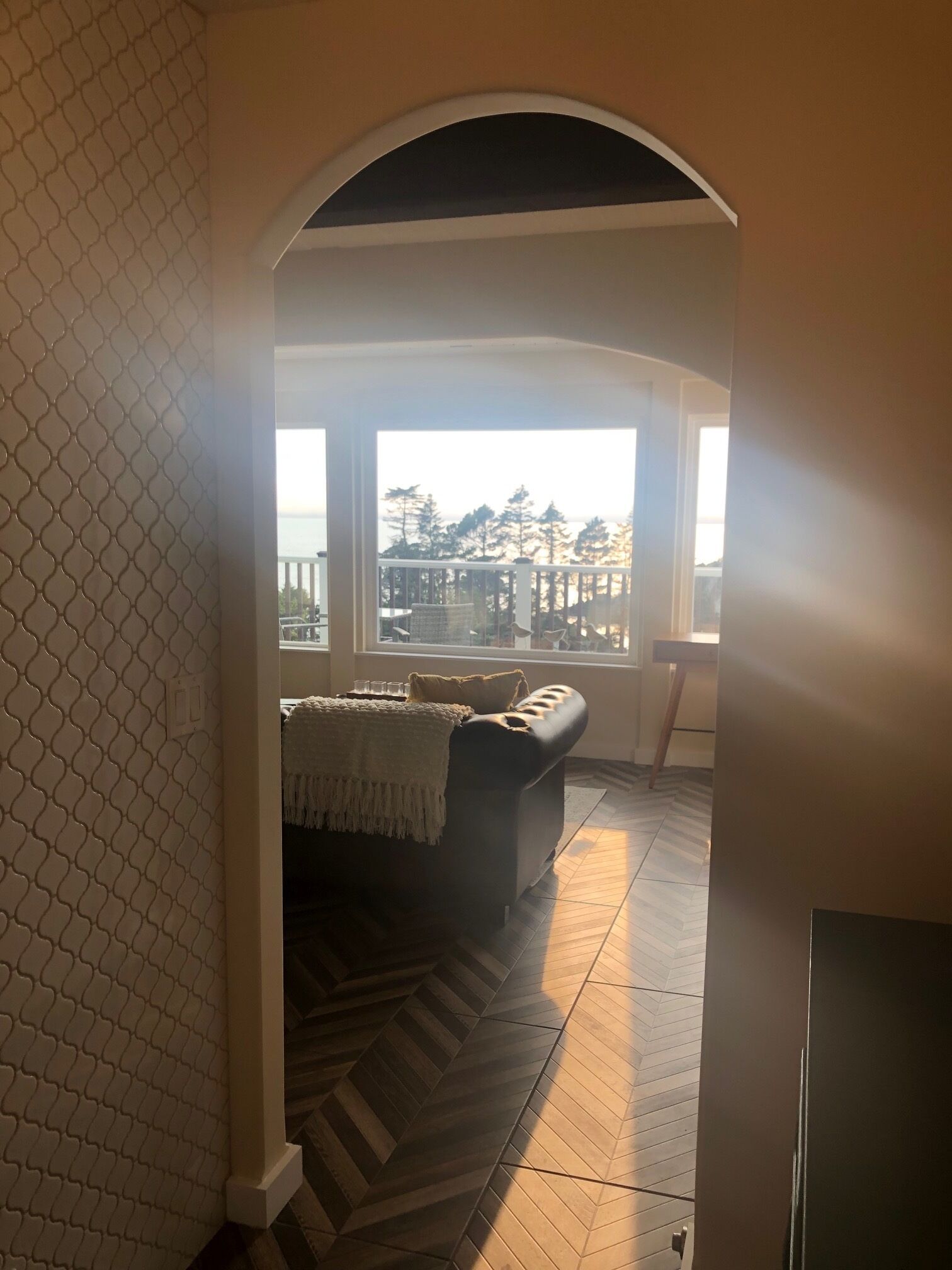 Ein Zimmer mit einem Fischgrätparkettboden, einem schwarzen Ledersofa und Blick auf einen Balkon mit Geländer und Bäumen.