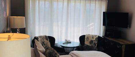 A bedroom with a bed, a desk, a chair, a lamp, a TV, and a window with curtains.