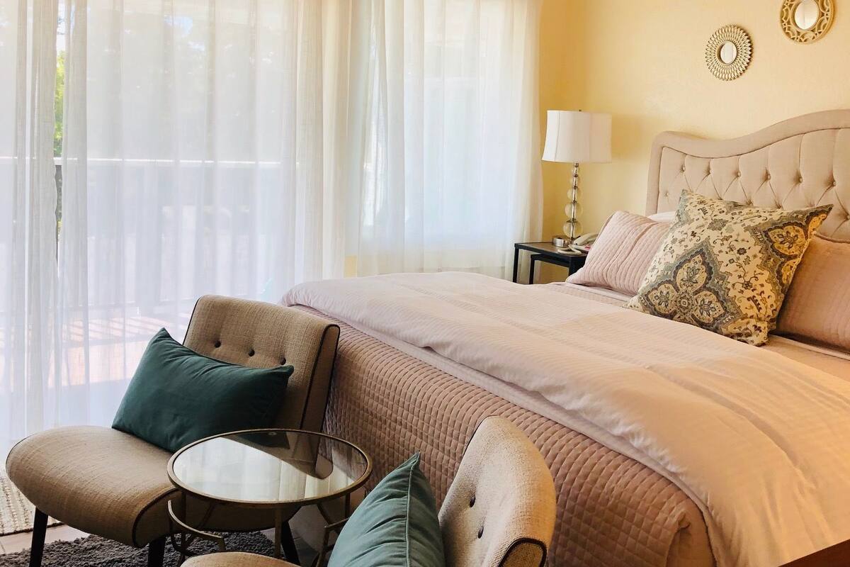A hotel room with a large bed, two armchairs, a small table, and a view of the outdoors through sheer curtains.