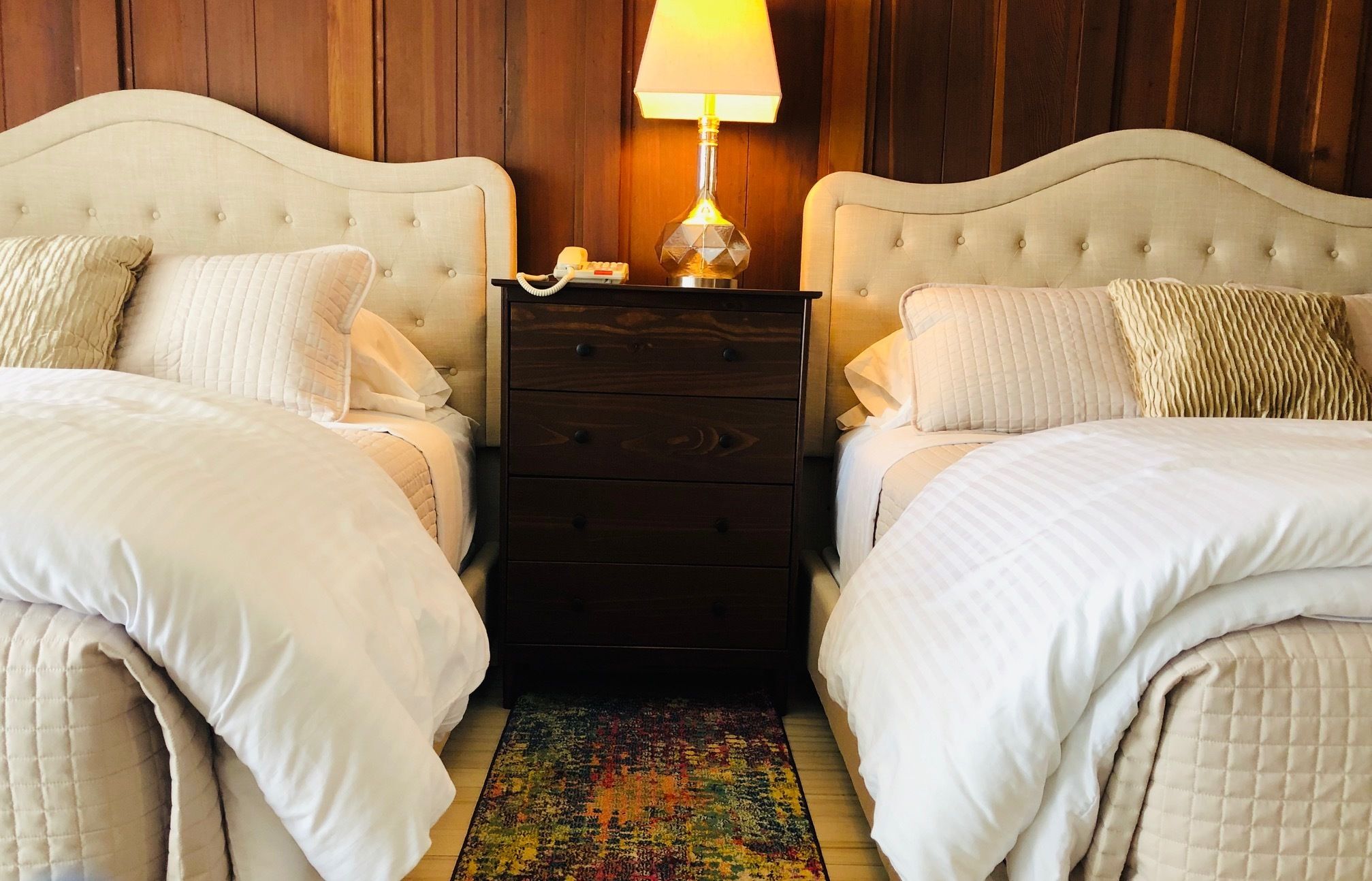 A hotel room with two beds, a nightstand with a lamp, and a colorful rug.