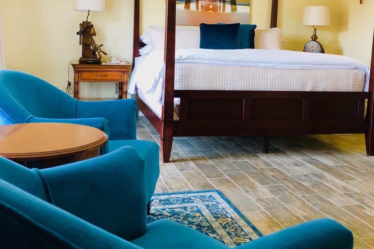 A four-poster bed with a view of a sunset, a blue armchair, a small round table, and a ship's wheel on the wall.