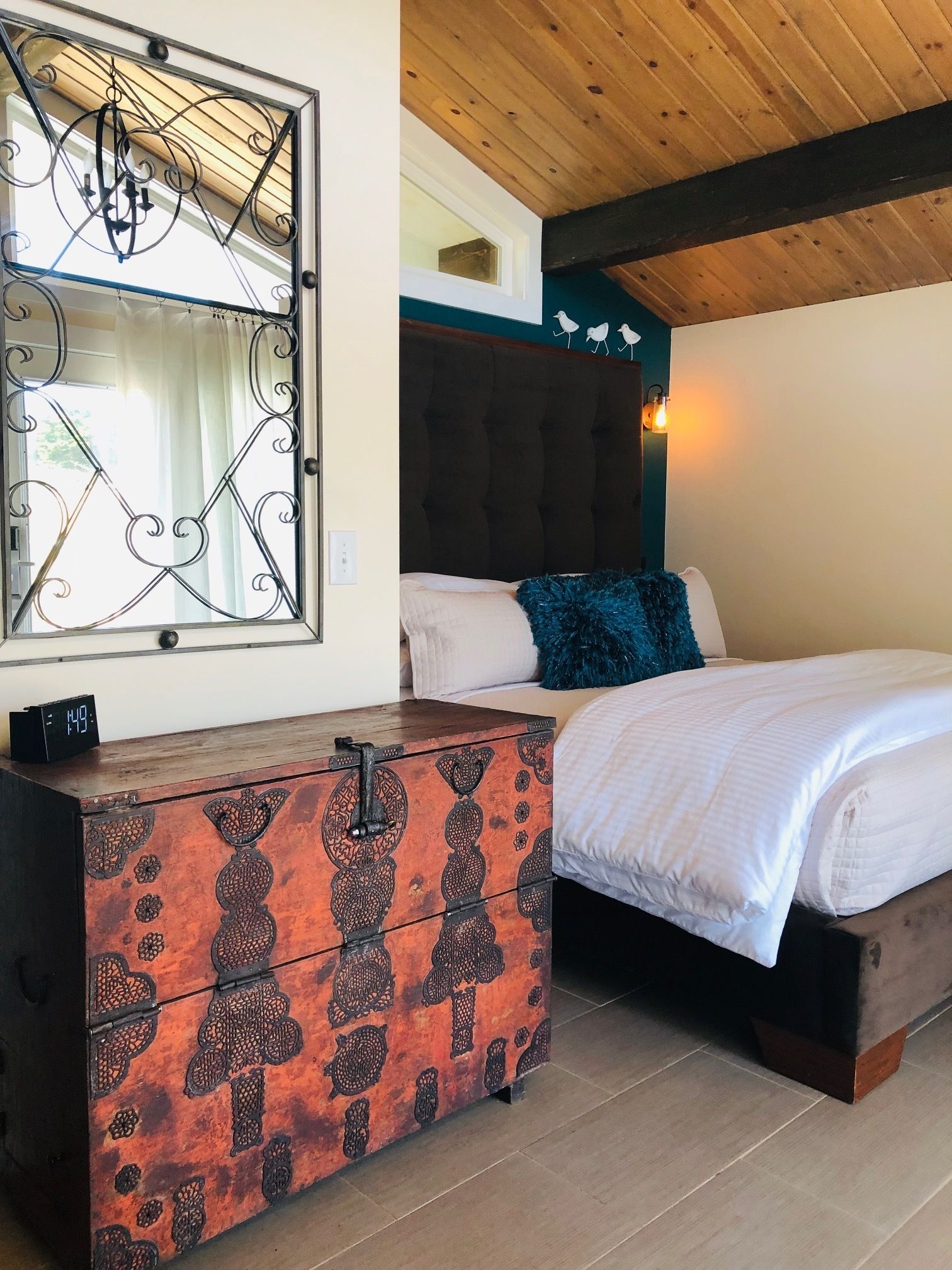 A bedroom with a wooden ceiling, a bed with white bedding, a decorative chest of drawers, and a wall-mounted light fixture.