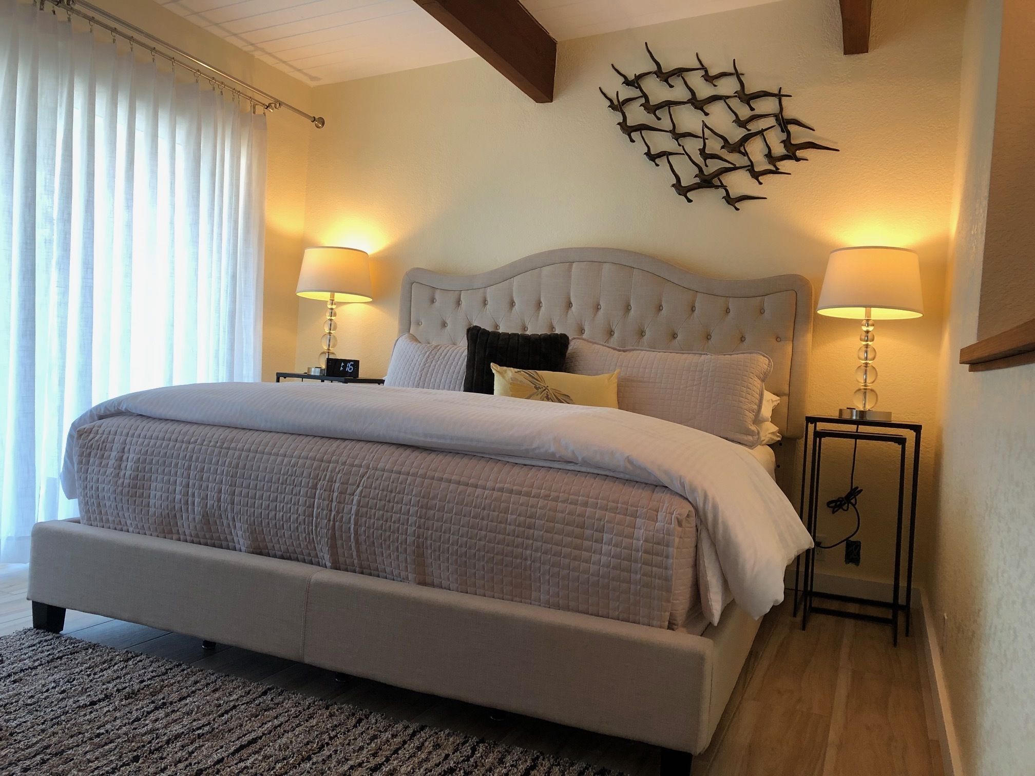 A large bed with a tufted headboard, two bedside lamps, and a wall-mounted abstract art piece.