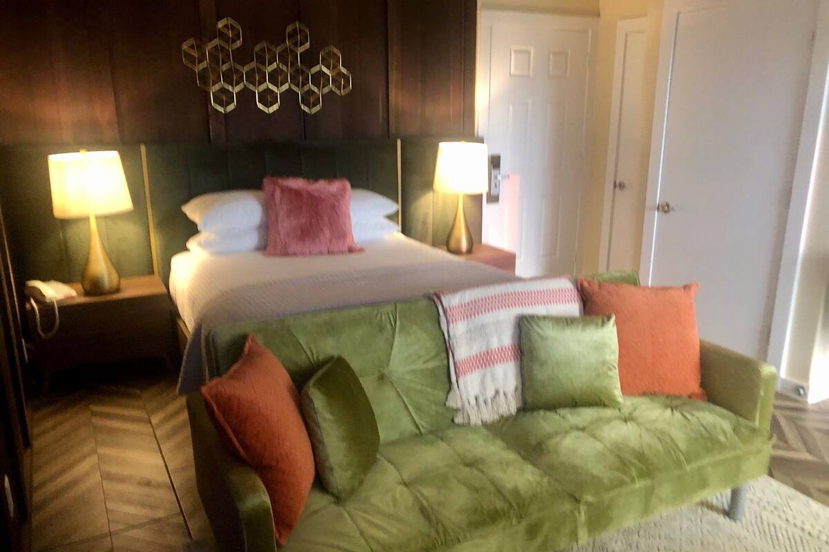 A hotel room with a green sofa, a bed with a pink pillow, two bedside lamps, and a geometric wall decoration.