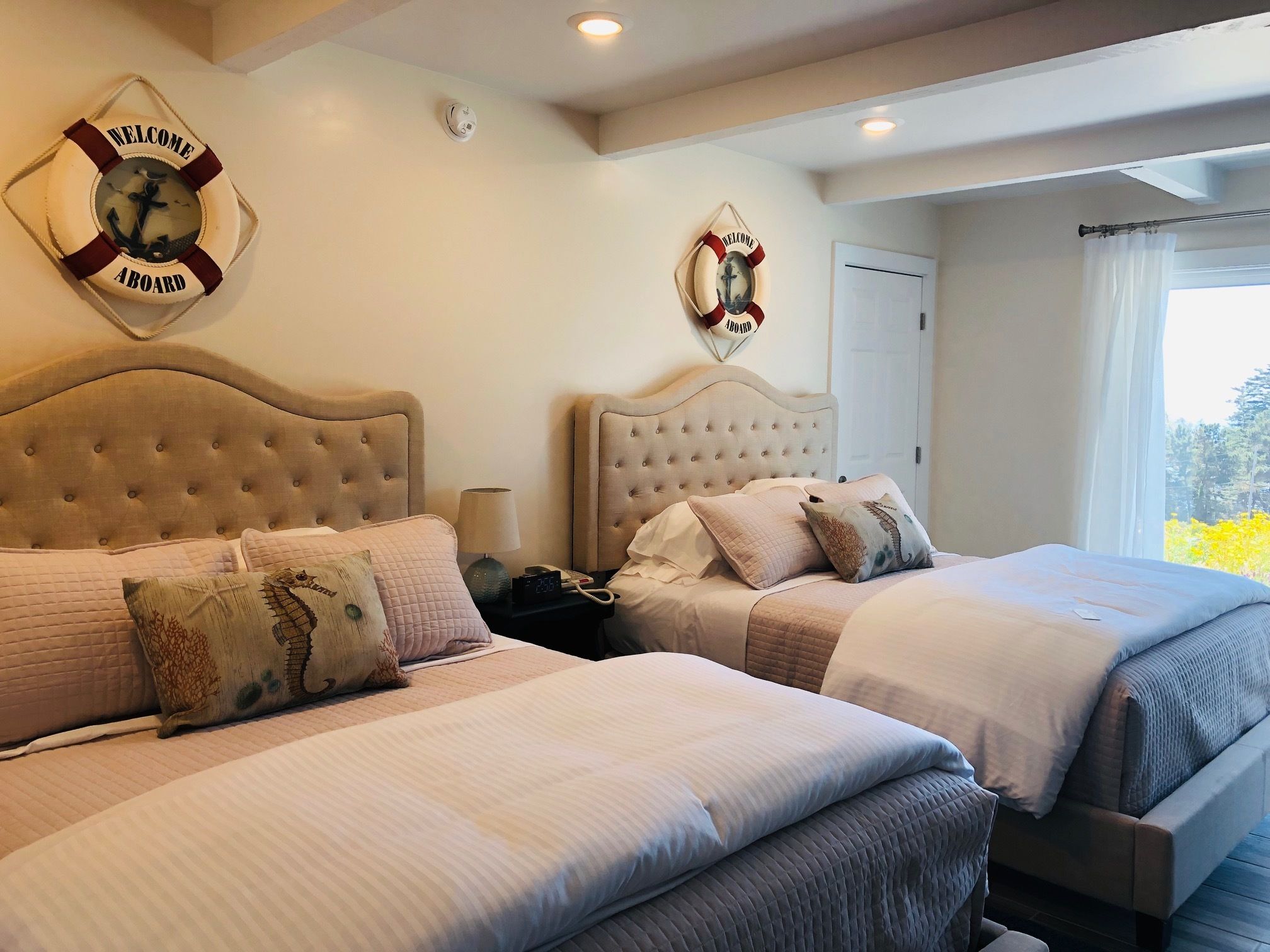A hotel room with two beds, each with a headboard and multiple pillows. There are wall-mounted clocks and a window with curtains.