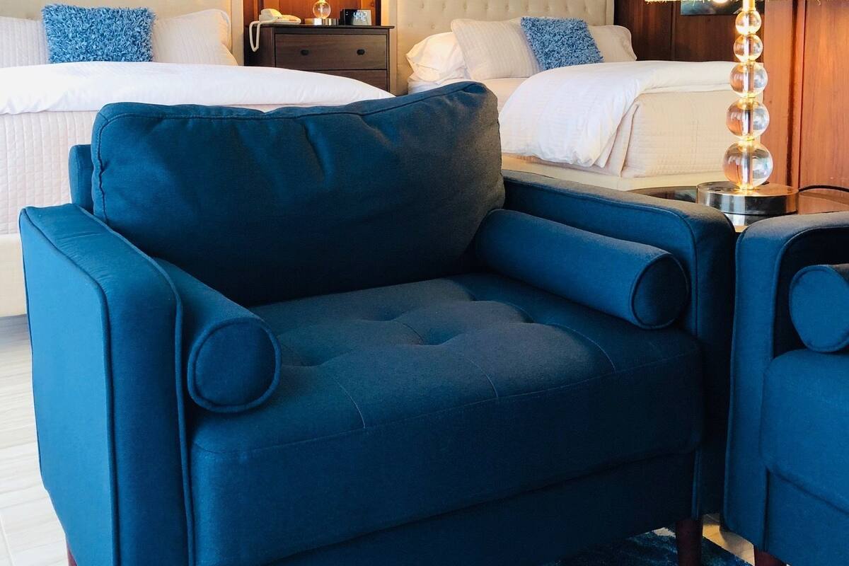 A hotel room with two beds, a blue armchair, a nightstand with a lamp, and wooden paneling on the walls.
