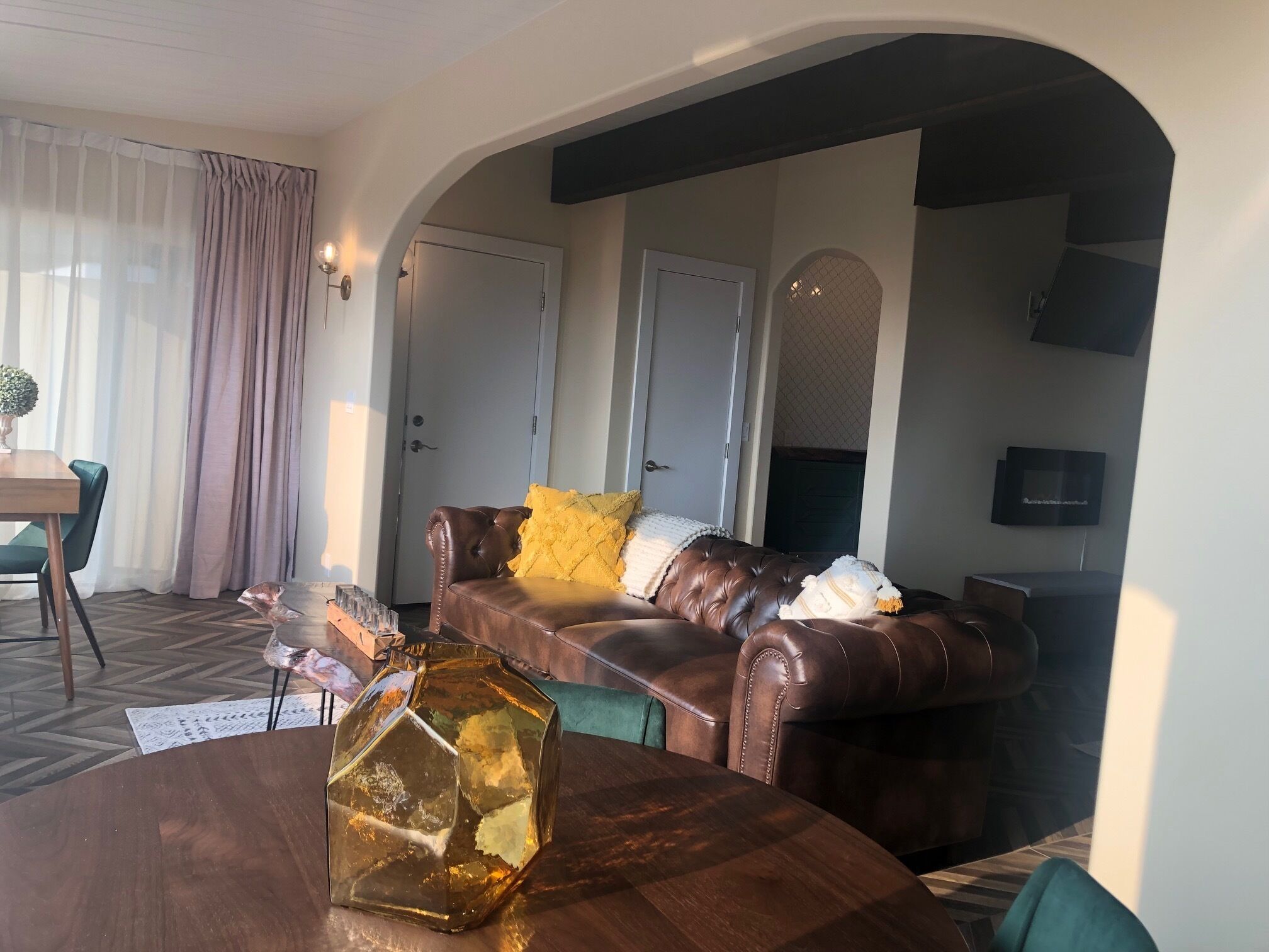 Ein Wohnzimmer mit einer braunen Ledersofa, einem runden Holztisch und einer sechseckigen Glasvase. Eine Tür mit einem Moskitonetz, ein an der Wand befestigtes Fernsehgerät und ein Fenster mit transparenten Vorhängen.