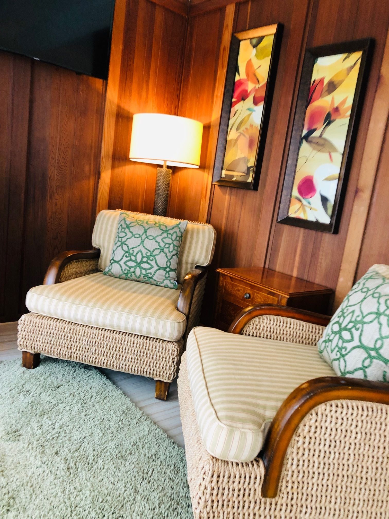 Um quarto aconchegante com duas poltronas de vime, uma mesinha de cabeceira de madeira, um abajur e duas pinturas florais abstratas na parede.