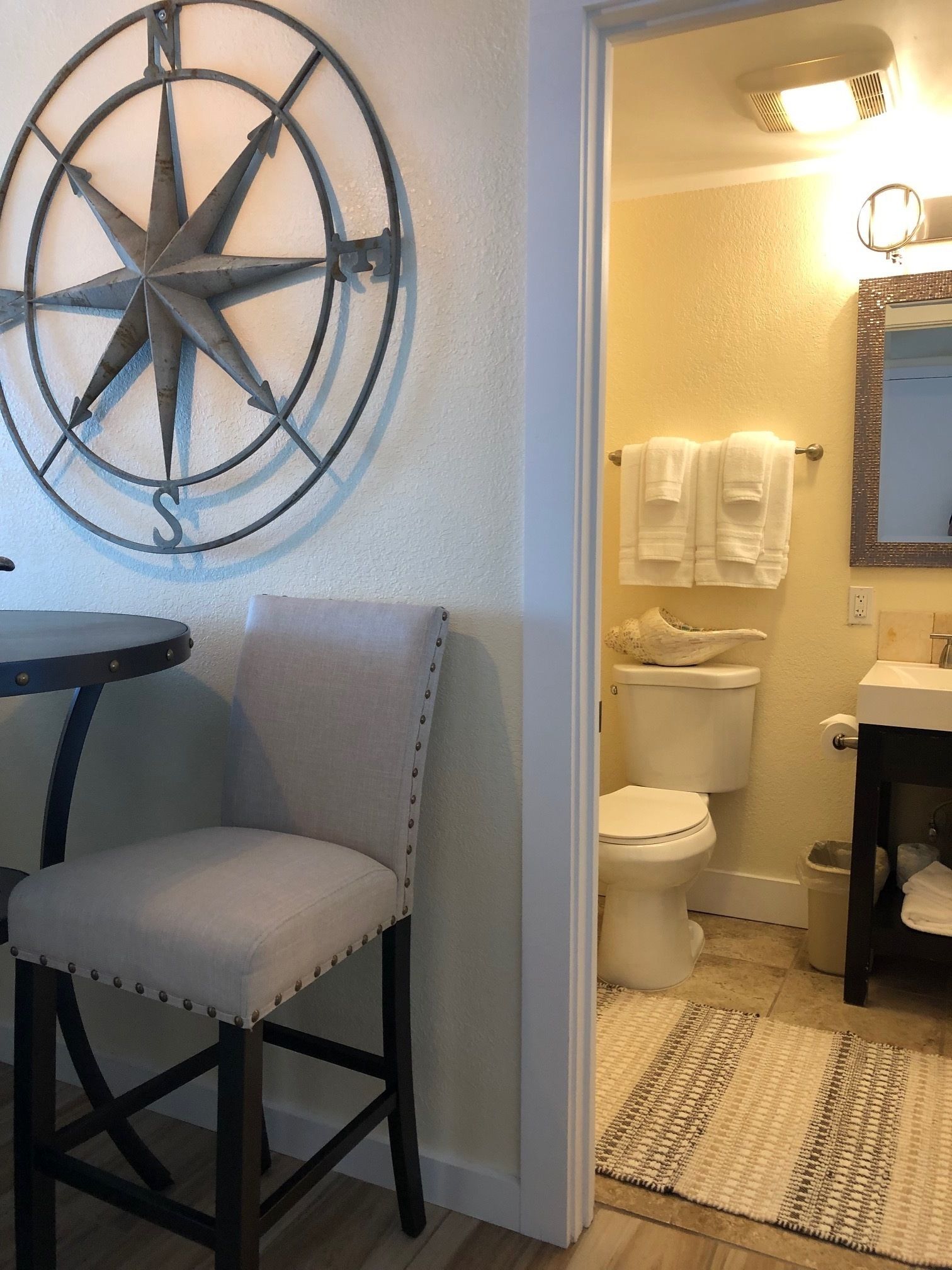 A bathroom with a toilet, sink, and mirror. There is a chair and a rug in the room.