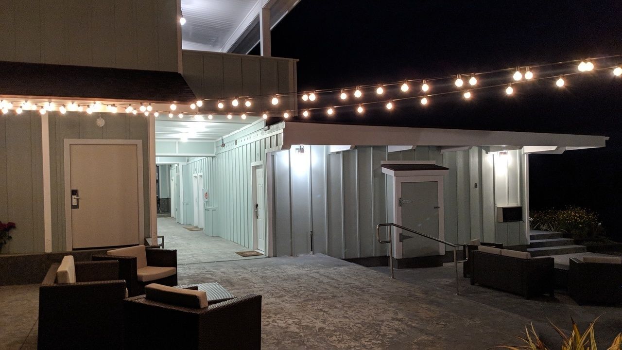Un patio al aire libre con luces colgantes, asientos exteriores y un corredor cubierto que lleva a un edificio con varias puertas.